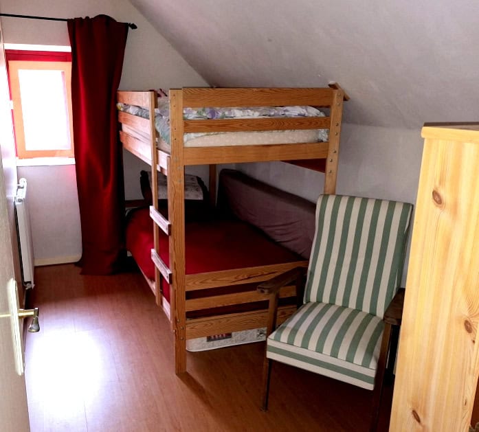 Attic bedroom featuring wooden bunk beds, a striped armchair, and a small window with red curtains.