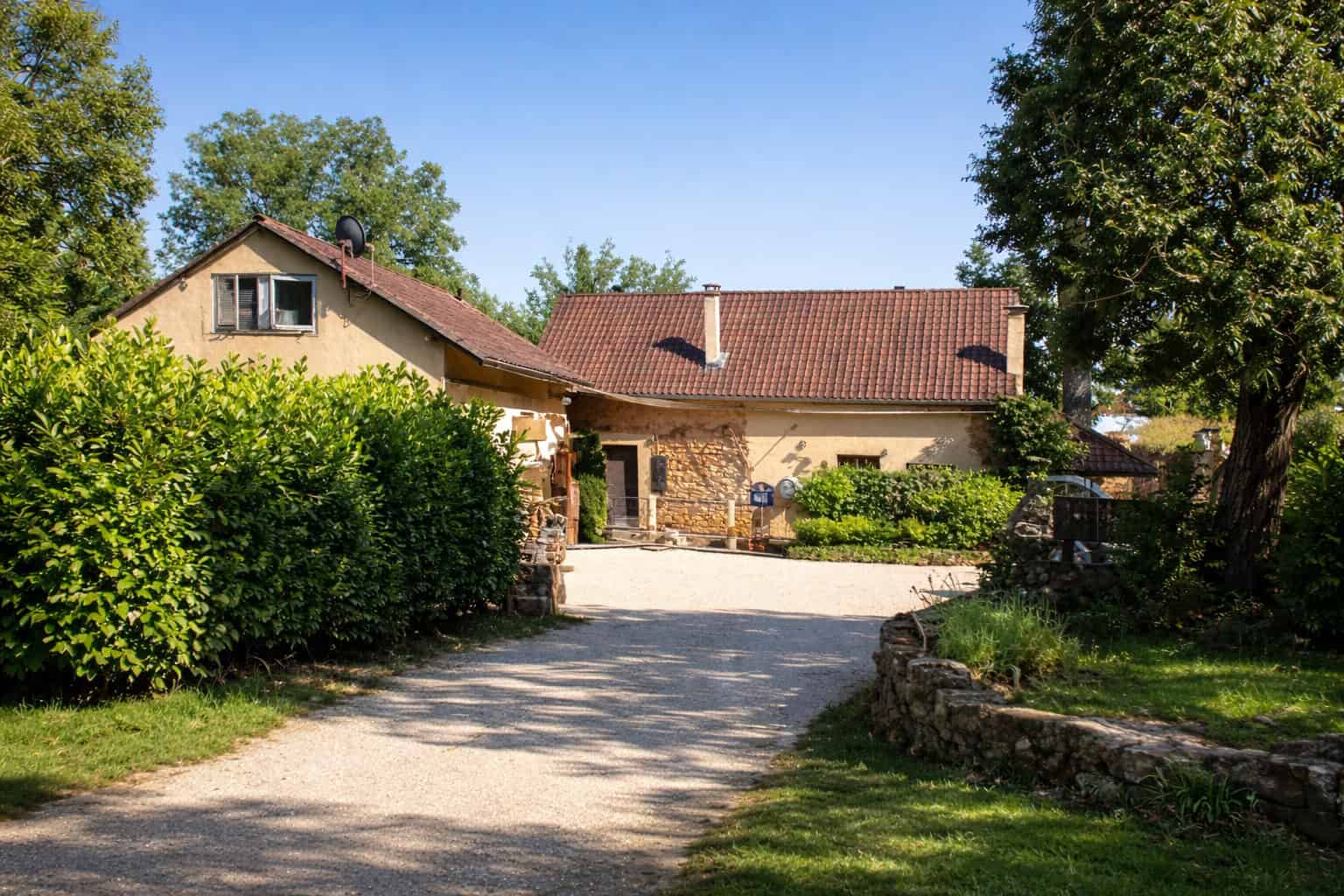 Rural house surrounded by greenery and trees, with a gravel driveway and traditional architecture.
