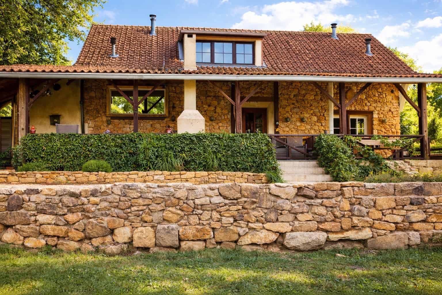 Stone cottage with red-tiled roof, surrounded by greenery and a stone wall.
