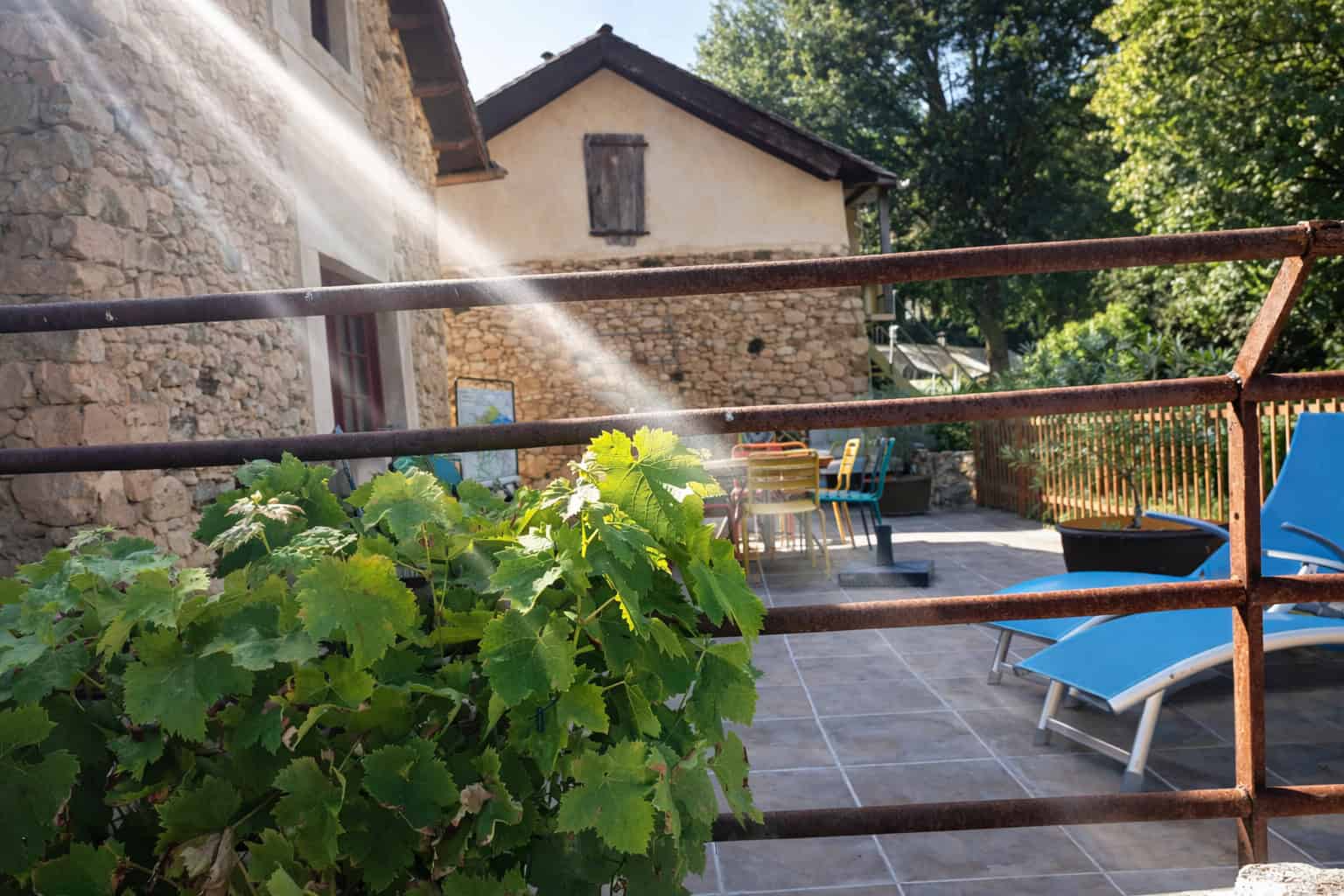 Outdoor terrace with lush green plants, sunlit seating area, and rustic stone buildings in the backg.